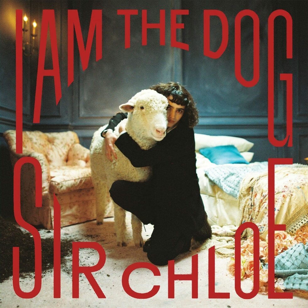 Album Art For I Am The Dog By Sir Chloe. Lead Singer Dana Foote Hugs A Fluffy White Sheep In A Dim, Disheveld Room.