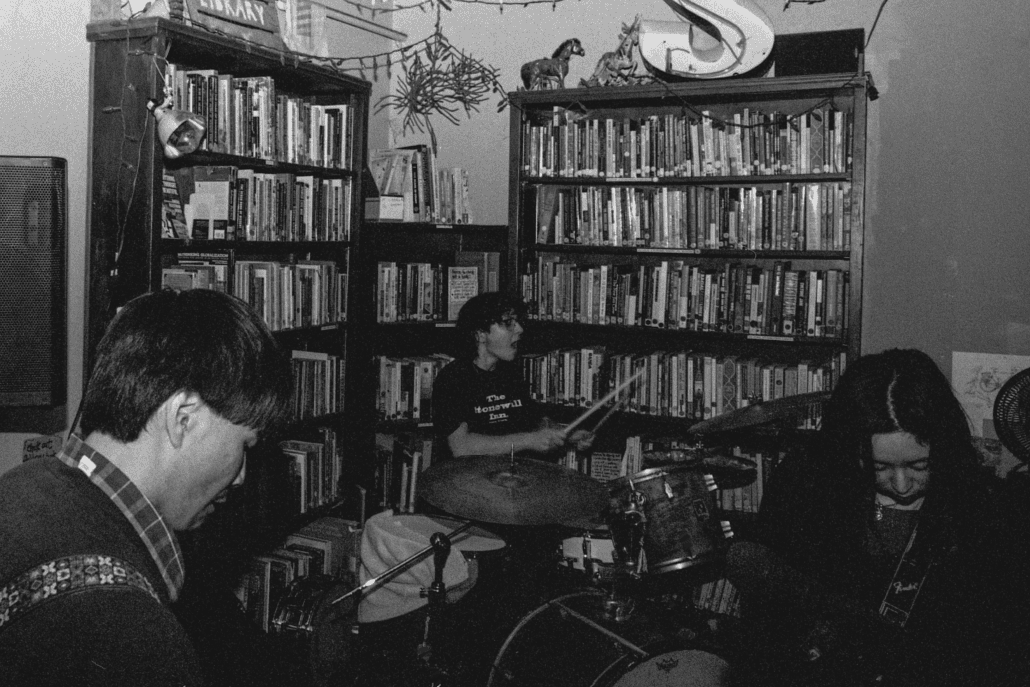 A Black-And-White Photo Shows Three Musicians Playing In A Cozy Room Lined With Bookshelves. One Plays Guitar, Another Drums, And The Third Is Partially Hidden, Possibly Playing Bass. String Lights And Animal Figurines Decorate The Space.